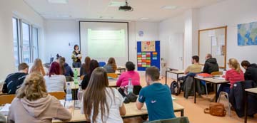 Klasse von hinten, Lehrerin mit Pinwand und Video unterrichtet